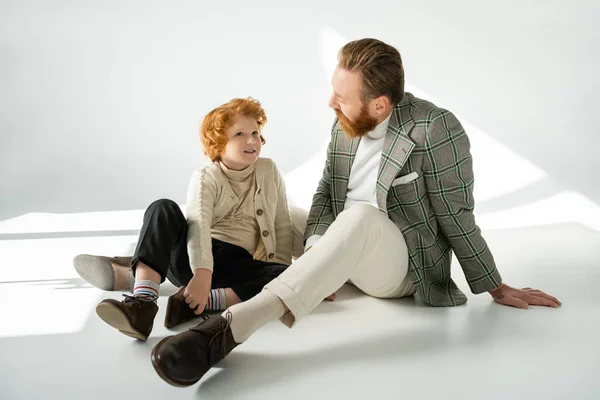 Улыбающийся рыжий парень в свитере разговаривает с бородатым отцом на сером фоне с солнечным светом — стоковое фото