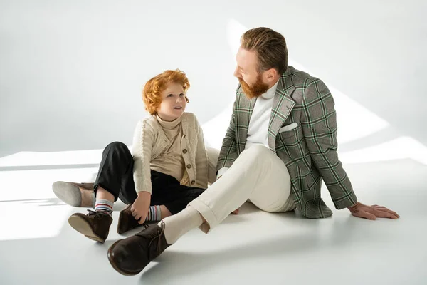 Souriant rousse garçon en pull parler à papa barbu sur fond gris avec la lumière du soleil — Photo de stock