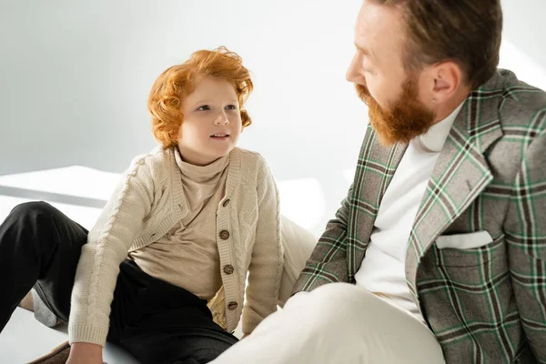 Smiling redhead boy in knitted jumper looking at blurred dad on grey background — Stock Photo