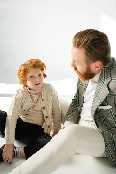 Red haired boy in jumper sitting near stylish dad on grey background with sunlight — Stock Photo