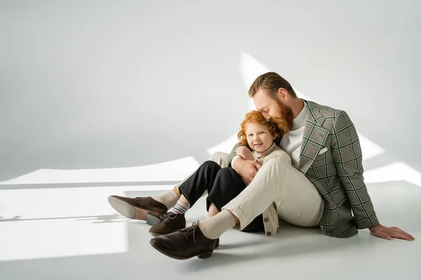 Sorrindo pai barbudo na jaqueta abraçando o filho de cabelos vermelhos enquanto sentado no fundo cinza com luz solar — Fotografia de Stock