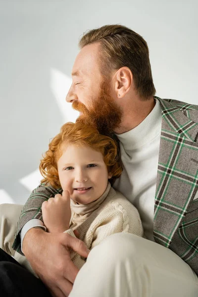 Бородатый мужчина в клетчатой куртке обнимает рыжеволосого сына на сером фоне со светом — Stock Photo