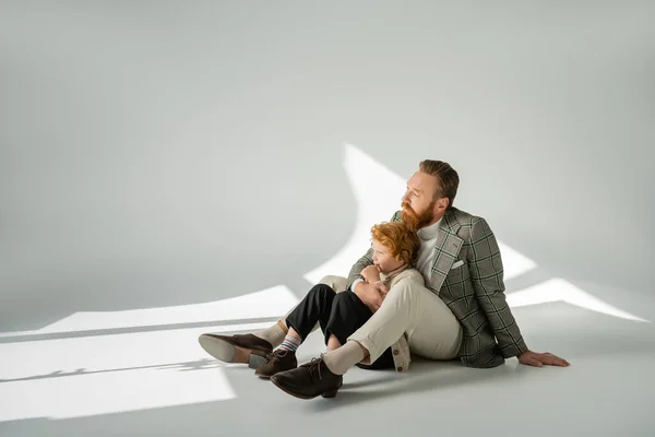 Hombre barbudo de moda abrazando al niño pelirrojo mientras está sentado sobre un fondo gris con luz solar - foto de stock