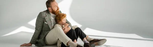 Man in jacket hugging redhead child while sitting on grey background with sunlight, banner — Stock Photo