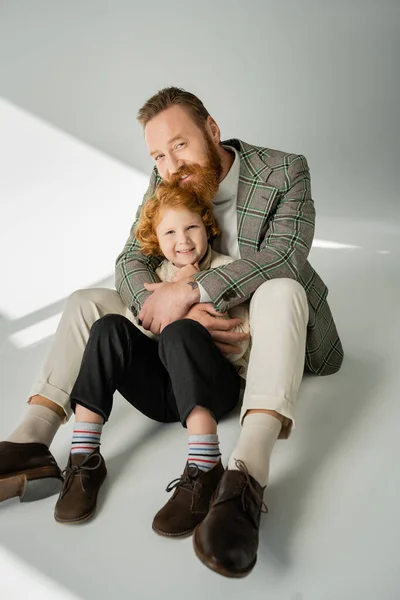 Trendy father hugging cheerful redhead child on grey background with sunlight — Stock Photo