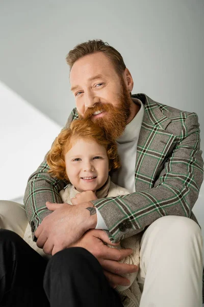 Portrait of man hugging red head son and looking at camera on grey background with light — Stock Photo