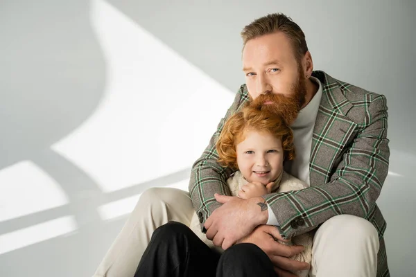 Bearded father hugging redhead son in knitted jumper on grey background with sunlight — Stock Photo