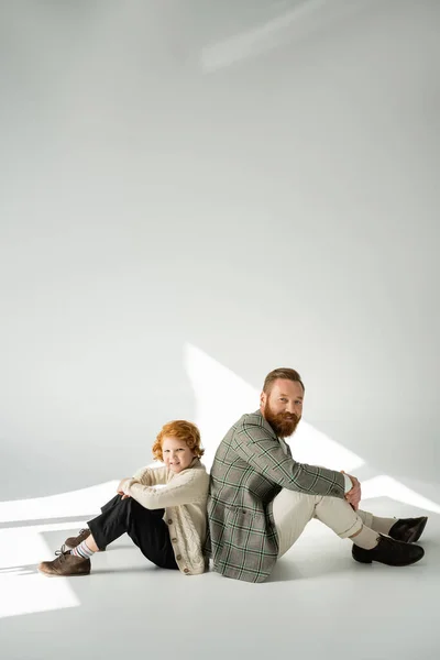 Stylish red haired kid and father sitting back to back on grey background with sunlight — Stock Photo