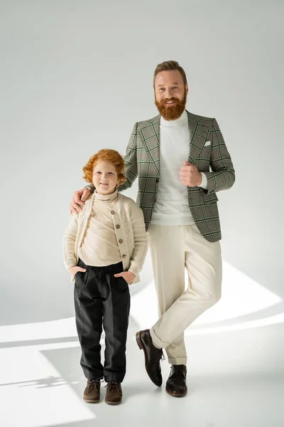 Père barbu à la mode et fils roux posant sur fond gris avec la lumière du soleil — Photo de stock