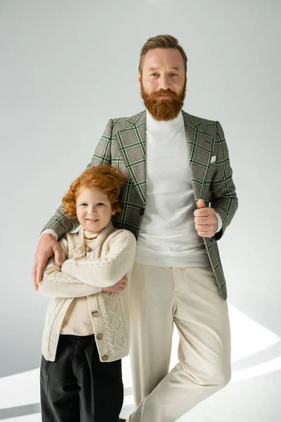 Fashionable man in jacket posing and hugging red haired son on grey background with natural light — Stock Photo