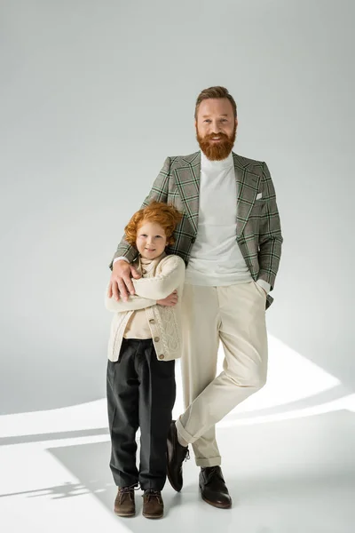 Volle Länge von stilvollen Vater und rothaarigen Sohn posiert und schaut in die Kamera auf grauem Hintergrund mit Licht — Stockfoto