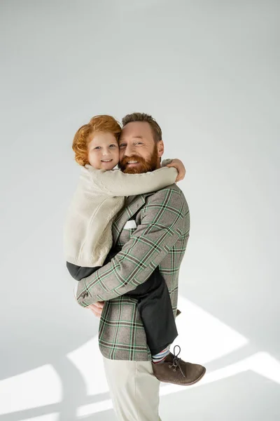 Happy bearded man in checkered jacket looking at camera while hugging son on grey background — Stock Photo