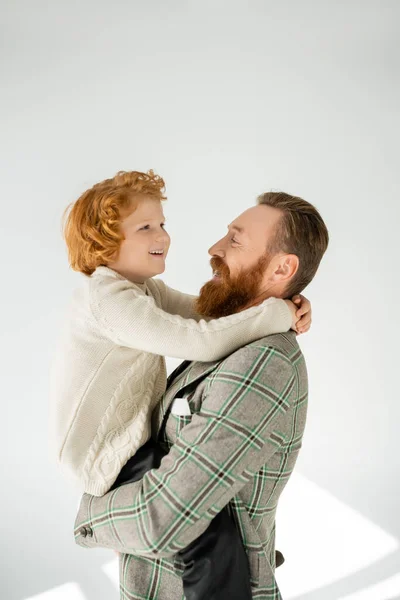 Трепетный отец в пиджаке обнимает и поднимает жизнерадостного рыжего сына на сером фоне — стоковое фото