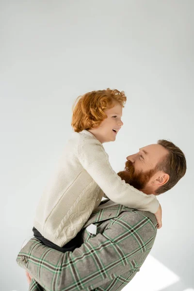 Cheerful bearded man lifting redhead son on grey background with sunlight — Stock Photo