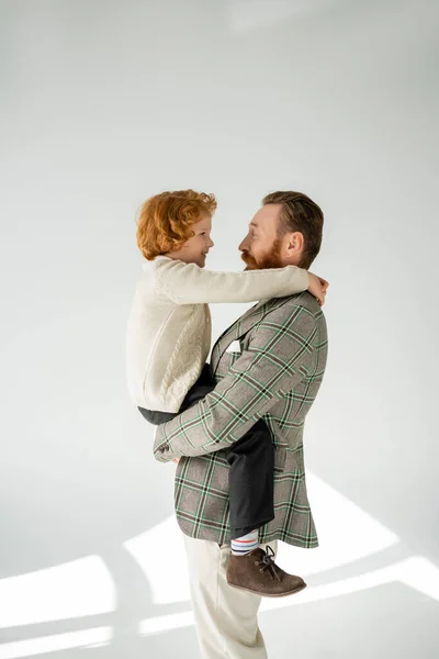 Side view of cheerful man in jacket hugging red haired son on grey background with sunlight — Stock Photo
