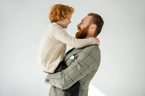 Vista lateral do pai positivo e barbudo que levanta o filho no jumper tricotado no fundo cinza — Fotografia de Stock
