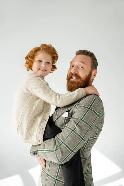 Smiling man in checkered jacket holding red haired son on grey background with sunlight — Stock Photo