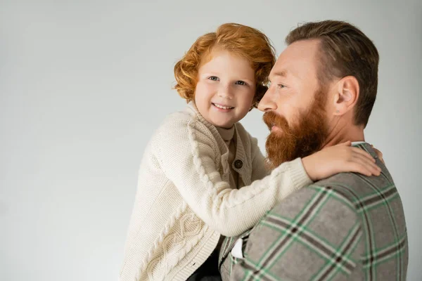 Menino de cabelos vermelhos em malha jumper abraçando pai barbudo e olhando para a câmera isolada em cinza — Fotografia de Stock