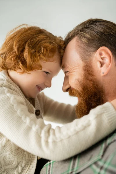 Niño de pelo rojo positivo abrazando a padre barbudo aislado en gris - foto de stock