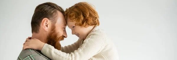 Vista laterale di positivo bambino dai capelli rossi in accogliente maglione abbracciando padre isolato su grigio, banner — Foto stock