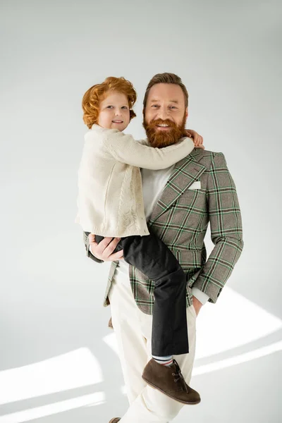 Stylish man posing and holding red haired son on grey background with sunlight — Stock Photo