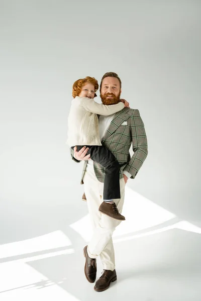 Full length of trendy bearded man holding red haired child on grey background with sunlight — Stock Photo