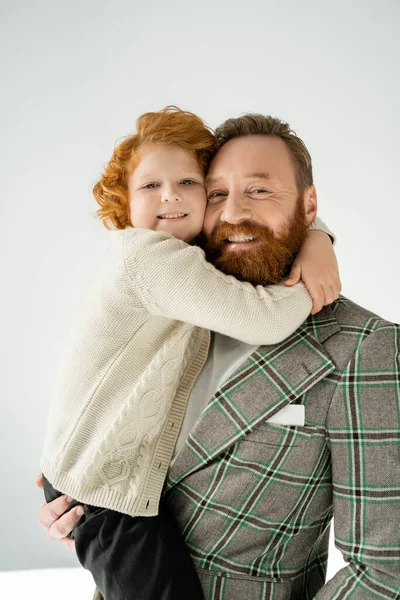 Red haired boy hugging stylish parent and looking at camera on grey background — Stock Photo