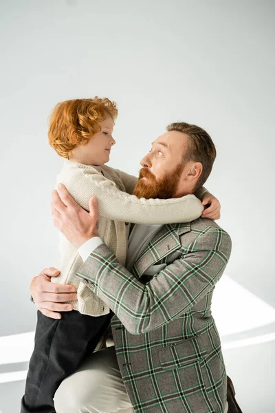 Excited man in jacket hugging smiling redhead son on grey background — Stock Photo