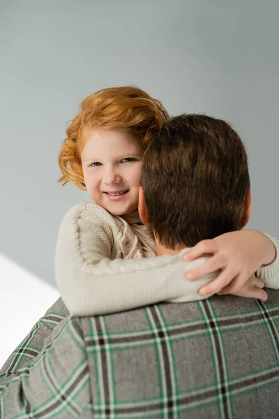 Positive Rotschopf-Kind in Strickpullover umarmt Papa auf grauem Hintergrund — Stockfoto