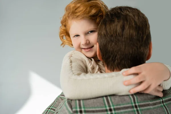 Cheerful red haired boy in knitted jumper hugging blurred dad on grey background — Stock Photo