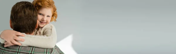 Garçon rousse souriant en pull tricoté embrassant parent flou sur fond gris, bannière — Photo de stock