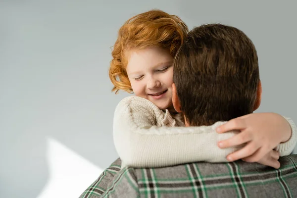 Garçon rousse en pull tricoté embrassant père et souriant sur fond gris — Photo de stock