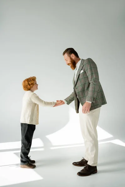 Side view of cheerful red haired boy in jumper clapping hand of bearded dad on grey background — Stock Photo