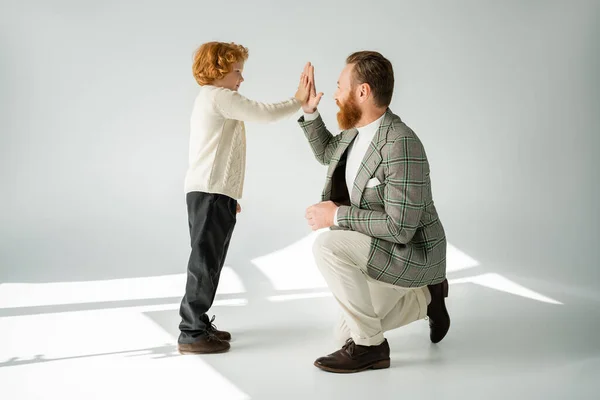 Seitenansicht des rothaarigen Jungen gibt High Five an stilvollen Vater auf grauem Hintergrund mit Licht — Stockfoto
