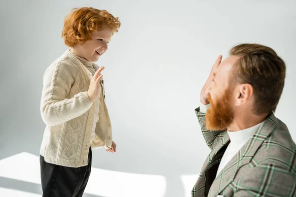 Enfant aux cheveux rouges souriant en pull tricoté donnant cinq à son père sur fond gris — Photo de stock