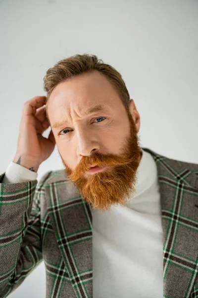 Портрет стильного и бородатого мужчины, трогающего волосы и смотрящего на камеру, изолированную на сером — стоковое фото