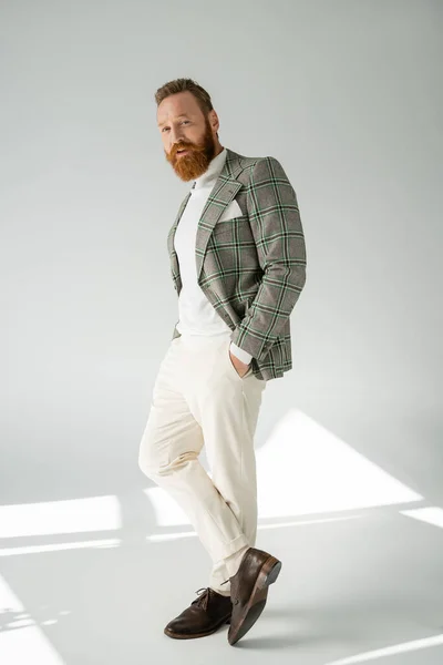 Toute la longueur de l'homme barbu élégant regardant la caméra sur fond gris avec la lumière du soleil — Photo de stock