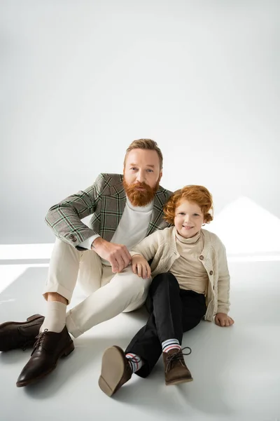 Бородатий батько і рудий волохатий син дивиться на камеру, сидячи на сірому фоні зі світлом — стокове фото