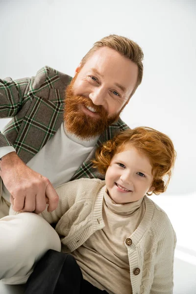 Portrait de père branché et garçon rousse souriant à la caméra sur fond gris avec lumière — Photo de stock