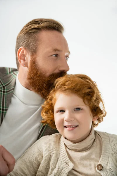 Бородатый мужчина целует улыбающегося рыжеволосого сына, изолированного от серых — стоковое фото