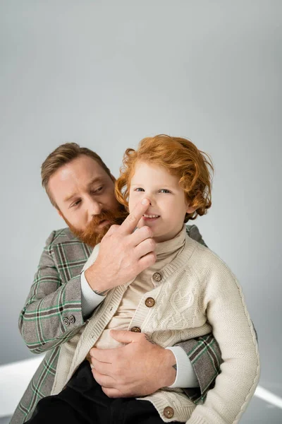 Бородатый мужчина в куртке касается носа рыжеволосого сына на сером фоне — стоковое фото