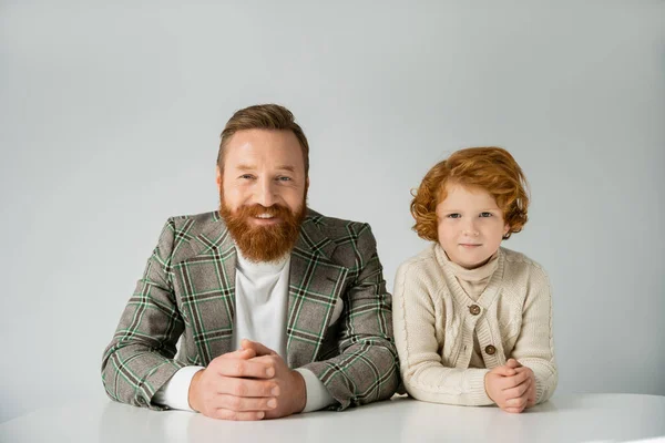 Redhead boy in knitted jumper looking at camera near smiling bearded dad and table isolated on grey — Stock Photo