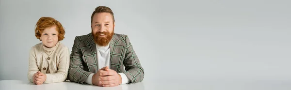 Homme barbu souriant en veste à carreaux regardant une caméra près d'un enfant roux isolé sur une bannière grise — Photo de stock