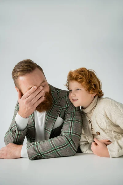 Cheerful red haired boy looking at stylish father near table isolated on grey — Stock Photo