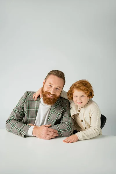 Smiling redhead boy hugging trendy father near table isolated on grey — Stock Photo