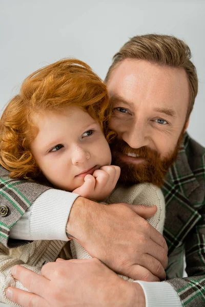 Бородатый мужчина обнимает рыжеволосого и веснушчатого сына, изолированного на сером — стоковое фото