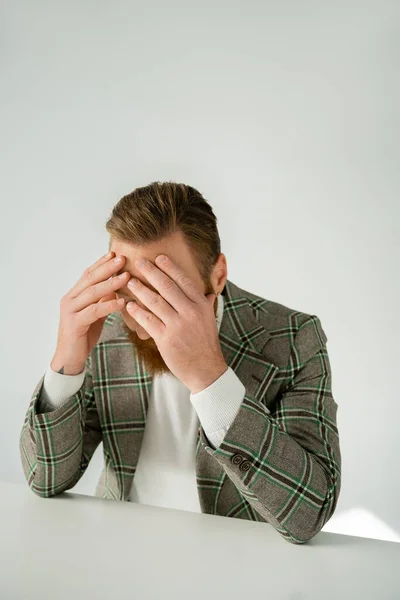 Bärtiger Mann in karierter Jacke berührt Stirn am Tisch vor grauem Hintergrund — Stockfoto