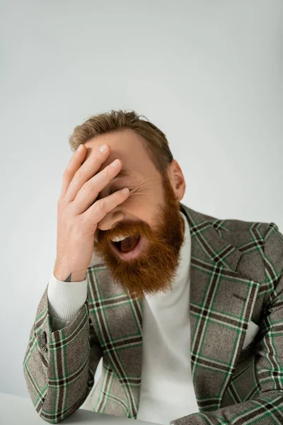 Стильный и бородатый мужчина в пиджаке смеется возле стола, изолированного на сером — стоковое фото