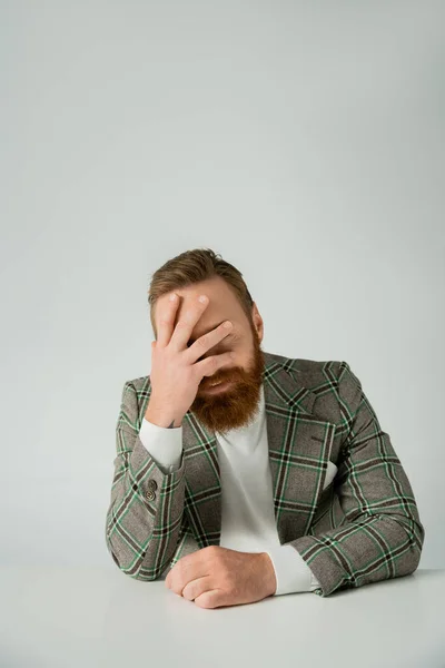 Улыбающийся бородатый мужчина в клетчатой куртке закрывает лицо рядом со столом, изолированным на сером — стоковое фото