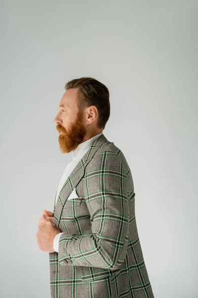 Vista lateral del hombre barbudo de moda ajustando solapas de chaqueta aisladas en gris - foto de stock
